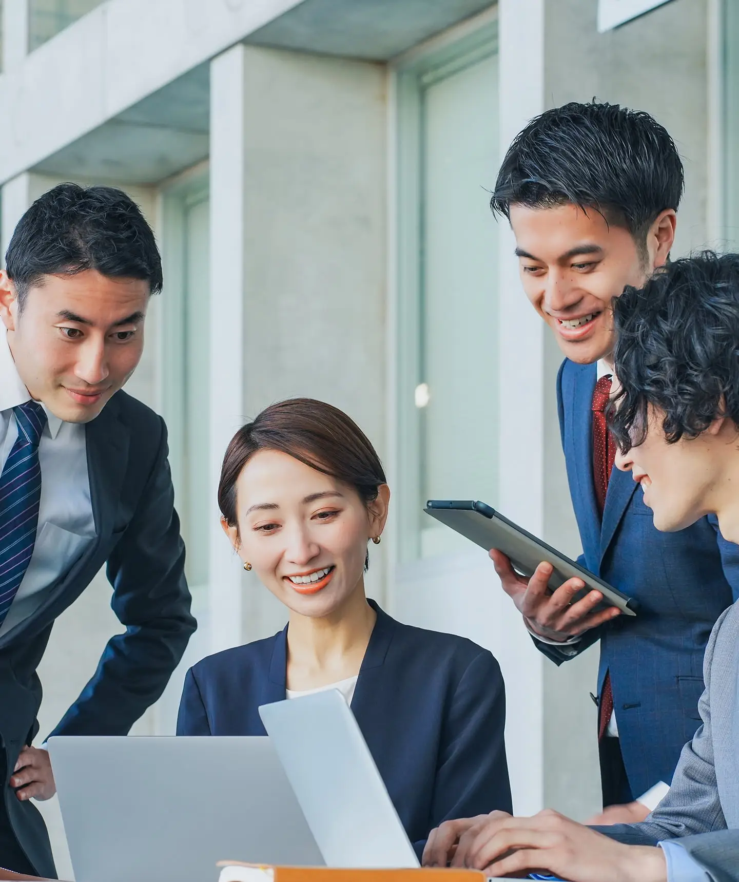 明るいオフィスで4人の若いビジネスパーソンがノートパソコンとスマートフォンを囲んで笑顔で協働している様子。チームワークと活気ある職場環境を表現