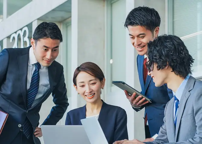 明るいオフィスで4人の若いビジネスパーソンがノートパソコンとスマートフォンを囲んで笑顔で協働している様子。チームワークと活気ある職場環境を表現