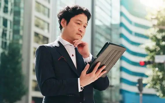 都市のビル群を背景に、スーツを着た男性がタブレットを見ながら顎に手を当てて考え込んでいる様子
