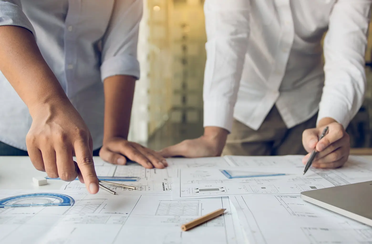 2人の人物が建築設計図を囲んで打ち合わせをしている様子。左側の人物は設計図上の特定の箇所を指差し、近くには分度器とコンパスが置かれている。右側の人物はペンを持ち、設計図に書き込みをしている。テーブル上にはノートパソコンや鉛筆も見える。