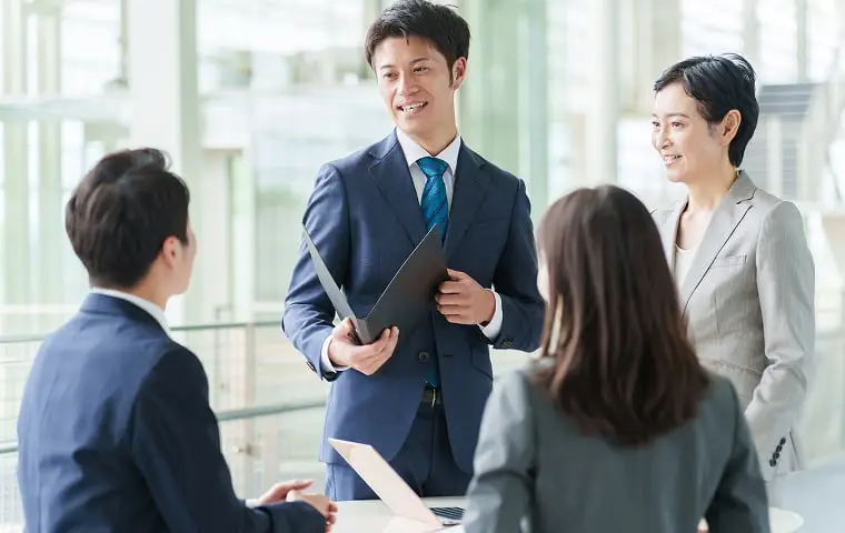 オフィスの会議で、紺のスーツを着た男性がファイルを持って立ち上がり、座っている同僚たちに説明している様子。グレーのスーツを着た女性も隣に立っている。