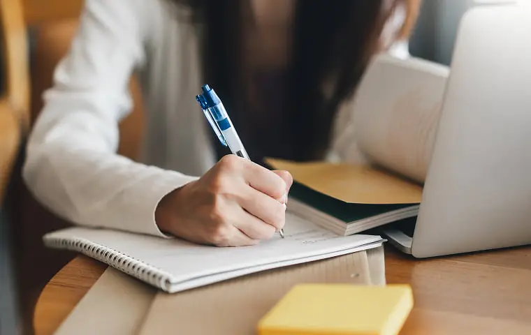 木製のデスクで、女性がノートパソコンの横でペンを使ってノートに書き込んでいる手元。デスク上にはノートパソコン、ノート、黄色い付箋が見える。