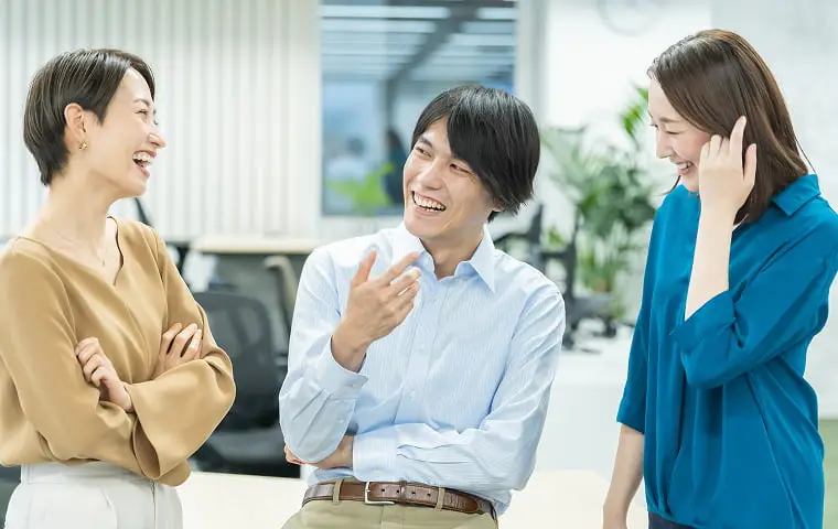 明るいオフィスで笑顔で会話する3人のビジネスパーソン（女性2人、男性1人）。ベージュのトップスを着た女性、白いシャツの男性、青いブラウスの女性が楽しそうに話している様子。