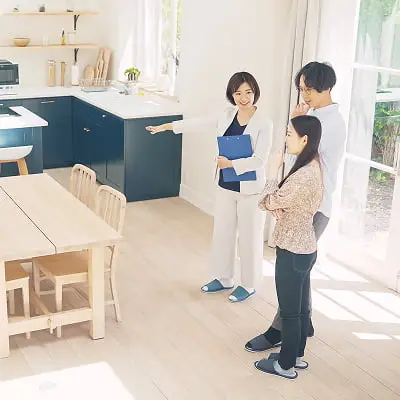 明るいモダンなキッチンダイニングで、青いエプロンを着た女性が資料を手に持ち、家族（男性と女の子）に説明している様子。住宅の内覧や不動産相談のシーン。