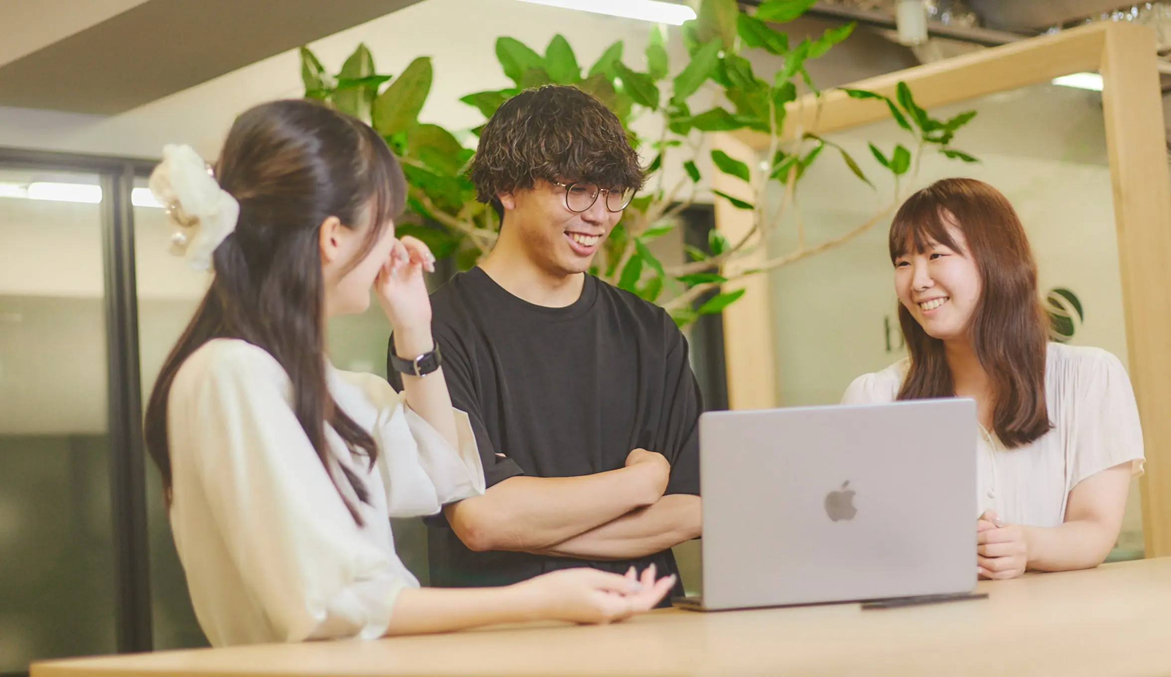 明るいオフィスで、ノートパソコンを囲んで3人のビジネスパーソンが笑顔で談笑している様子。左の白いブラウスの女性は髪に飾りをつけ、中央の黒いTシャツの男性は眼鏡をかけ腕を組み、右の白いトップスの女性はMacBookの前に座っている。背景には観葉植物が見える