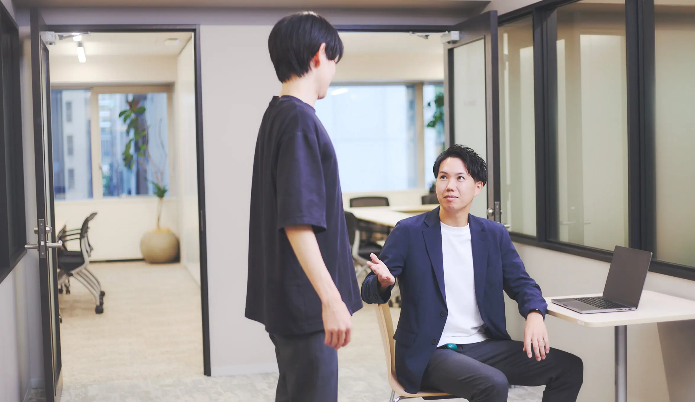 明るいモダンなオフィスで、2人の男性が会話している様子。左の男性は黒いTシャツとパンツを着用し、右の男性は白いTシャツの上に紺色のブレザーを着て椅子に座っている。座っている男性は左手を広げて何かを説明しているようで、立っている男性は彼の方を向いている。背景にはガラス張りの会議室や窓、観葉植物が見える