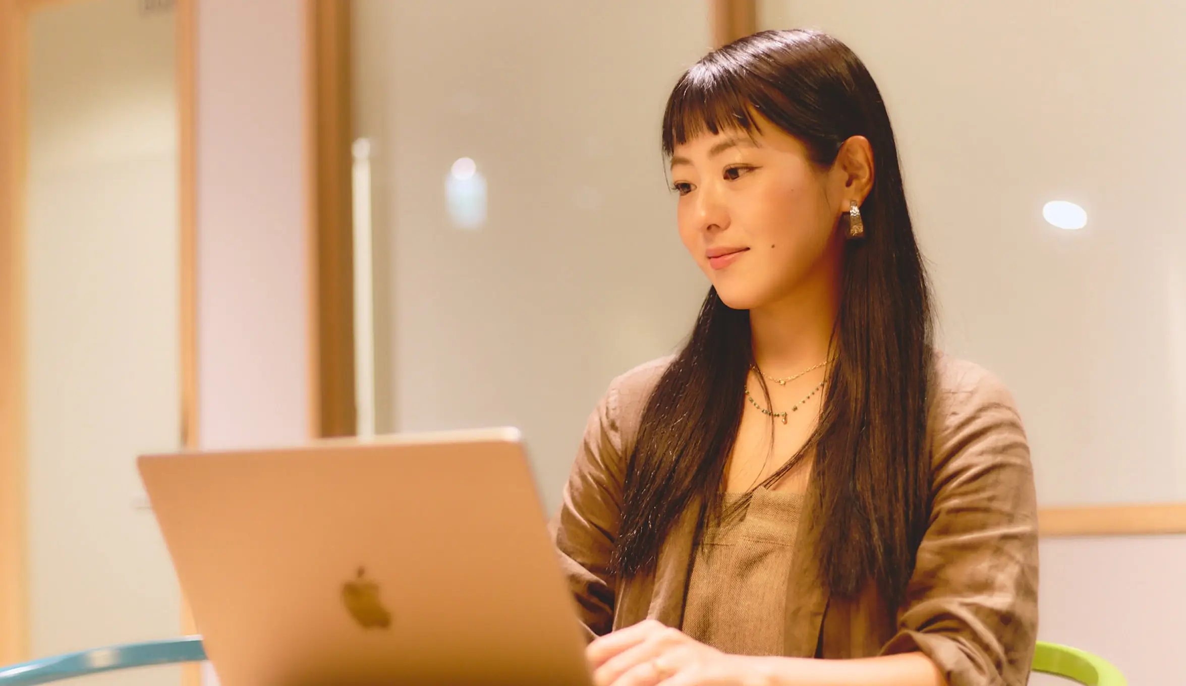 長い黒髪の女性が、明るいオフィスでシルバーのノートパソコンに向かって作業している様子。女性は茶色の重ね着をしており、繊細なネックレスとイヤリングを着用している。穏やかな表情で画面を見つめており、背景は暖色系の壁がぼやけて見える