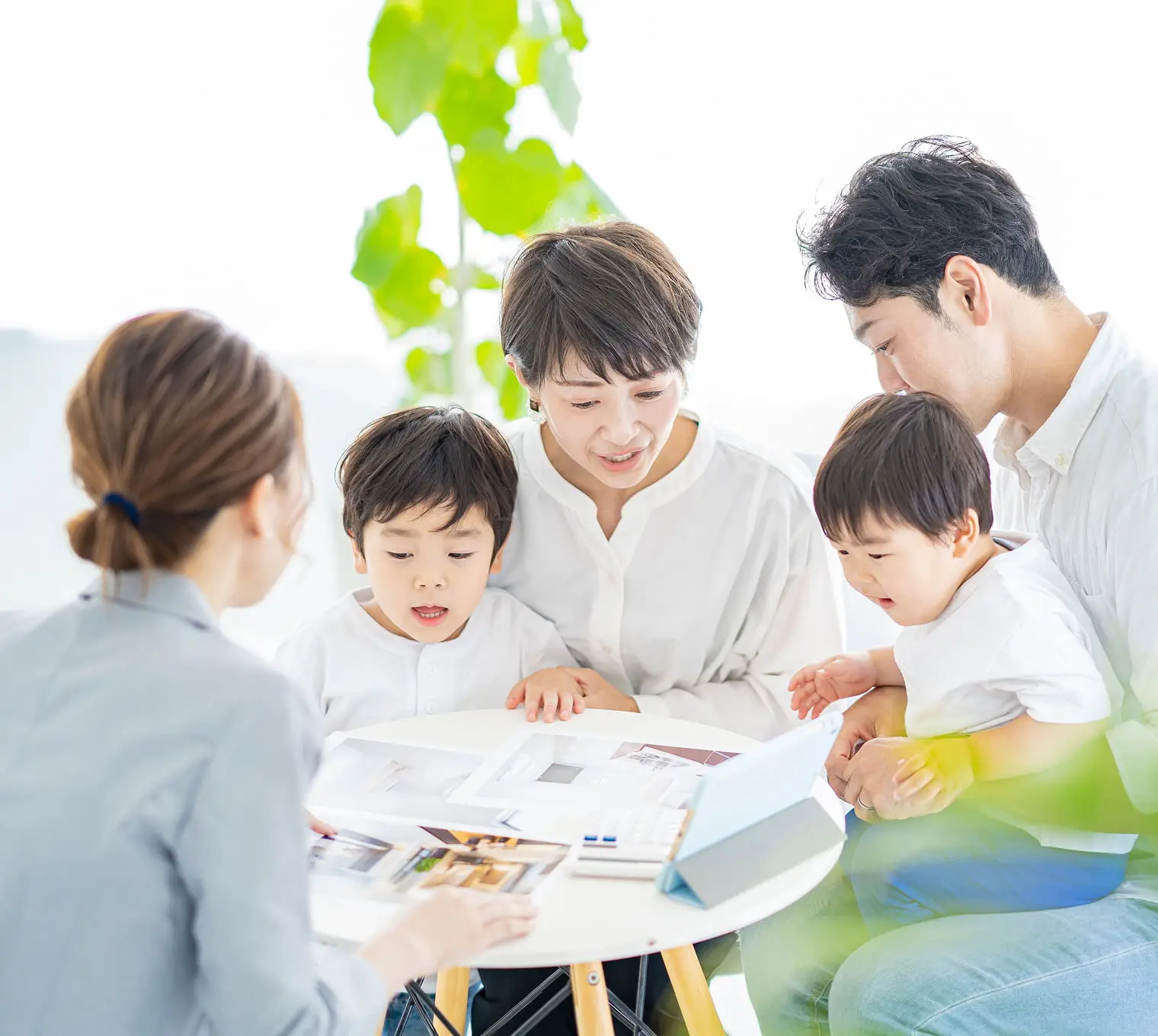 明るい室内で、4人家族（父親、母親、2人の子供）が女性スタッフと資料を囲んで相談している様子。テーブル上には住宅関連の資料やカタログが広げられ、観葉植物も見える。マイホーム購入や不動産の相談風景。