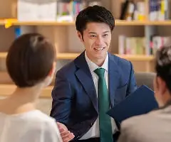 緑のネクタイをしたスーツ姿の男性が笑顔で相手と対面して話している様子。背景には本棚があり、コンサルティングや相談のシーン。