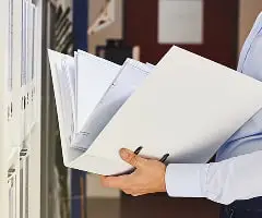 白いシャツを着たビジネスパーソンが書類やファイルを持っている様子。背景にはオフィスの棚が見える。