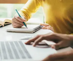 黄色いセーターを着た人が青いペンでノートに書き込んでいる手元。デスク上にはノートパソコンと書類が広げられている。