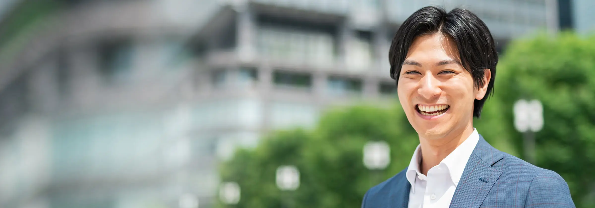 都市の緑豊かな環境で大きな笑顔を見せるスーツ姿の若い男性。爽やかで親しみやすい表情で、背景にはビルと緑が広がる。