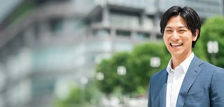 都市の緑豊かな環境で大きな笑顔を見せるスーツ姿の若い男性。爽やかで親しみやすい表情で、背景にはビルと緑が広がる。