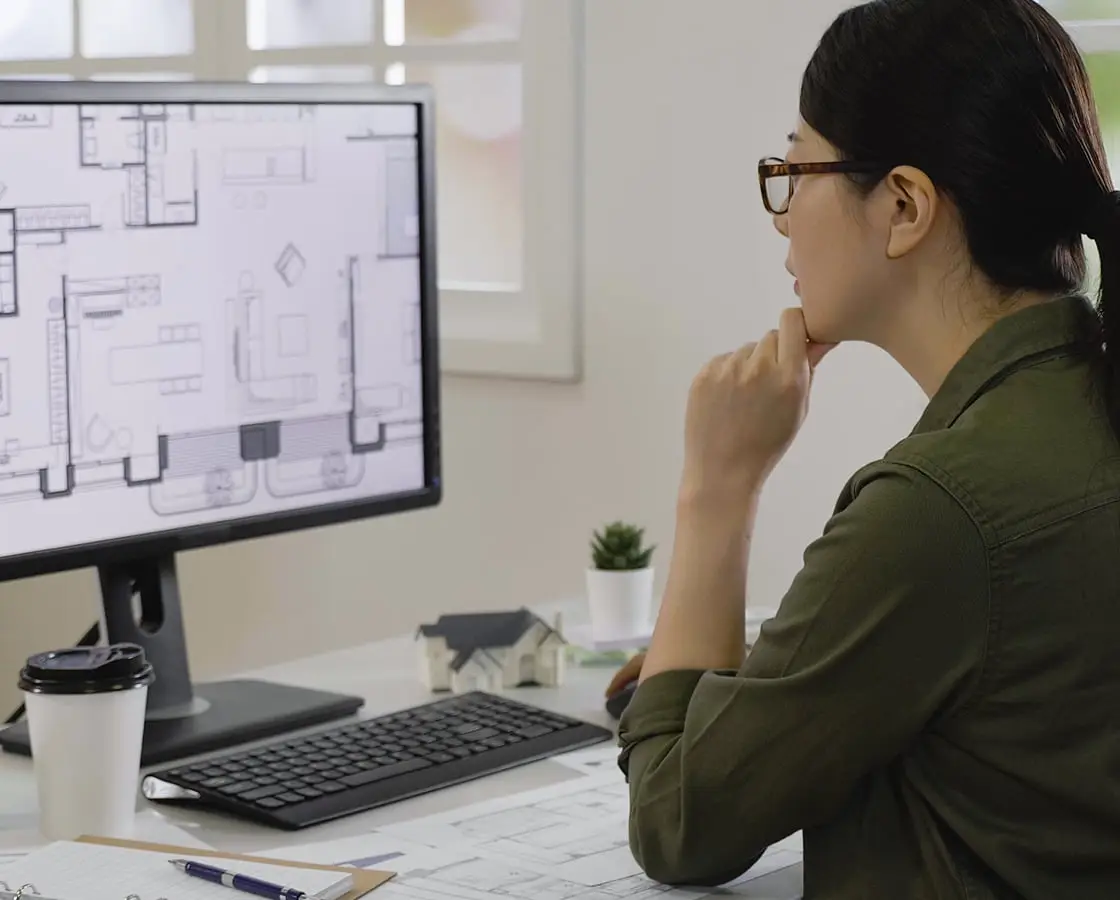 緑色のシャツを着た眼鏡の女性が、デスクトップモニターに表示された建築図面やフロアプランを見ながら顎に手を当てて検討している様子。デスク上にはコーヒーカップ、設計図面、ペンが置かれている。