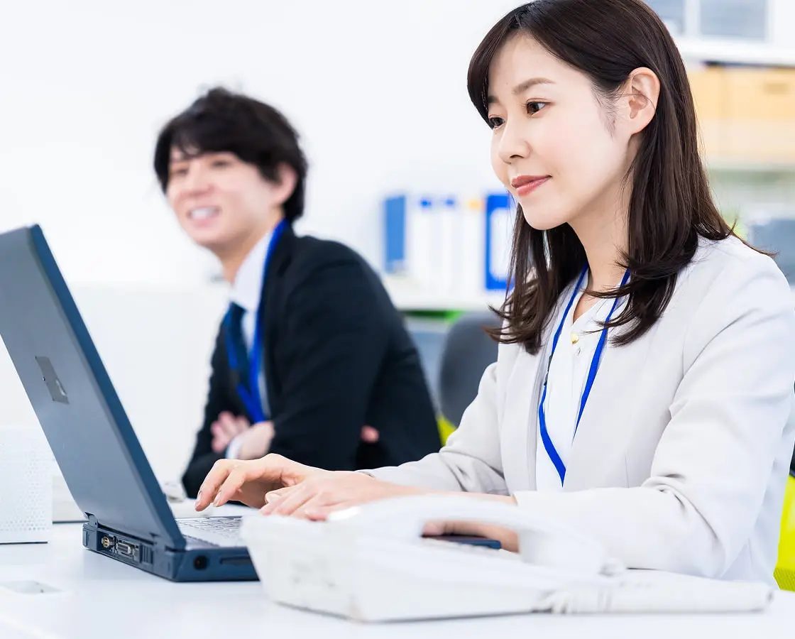 明るいオフィスで青いストラップを付けた女性がノートパソコンで作業している様子。背景には黒いスーツを着た男性も見える。
