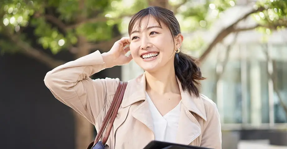 ベージュのトレンチコートを着た女性が屋外で笑顔で空を見上げている様子。手にスマートフォンと資料を持ち、緑豊かな背景が見える。