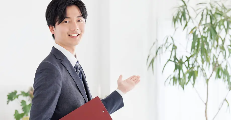 グレーのスーツを着た笑顔の若い男性が赤いファイルを持ち、手を差し出しながら案内している様子。背景には観葉植物が見える。