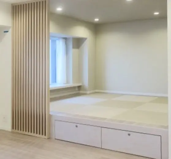 木製の縦格子パーテーションで仕切られたモダンな多目的ルーム。奥には白いカーテンのかかった窓とベンチがあり、手前には引き出し収納付きの小上がりが設けられ、市松模様の畳が敷かれている。手前の床は明るい木目調のフローリング。