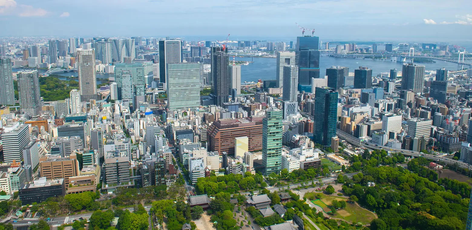 東京の都市景観の空撮。高層ビル群、東京湾、そして緑豊かな公園や伝統的な寺院建築が広がる様子