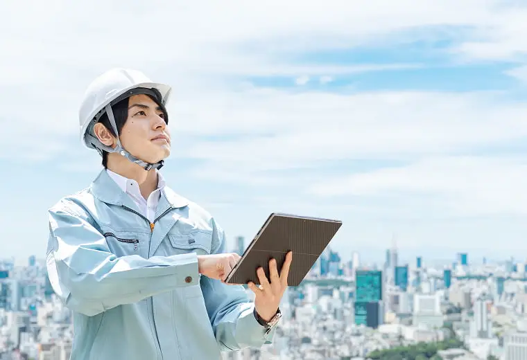 都市のビル群を背景に、ヘルメットを被りタブレットを操作しながら空を見上げる建設・エンジニアリング関係者