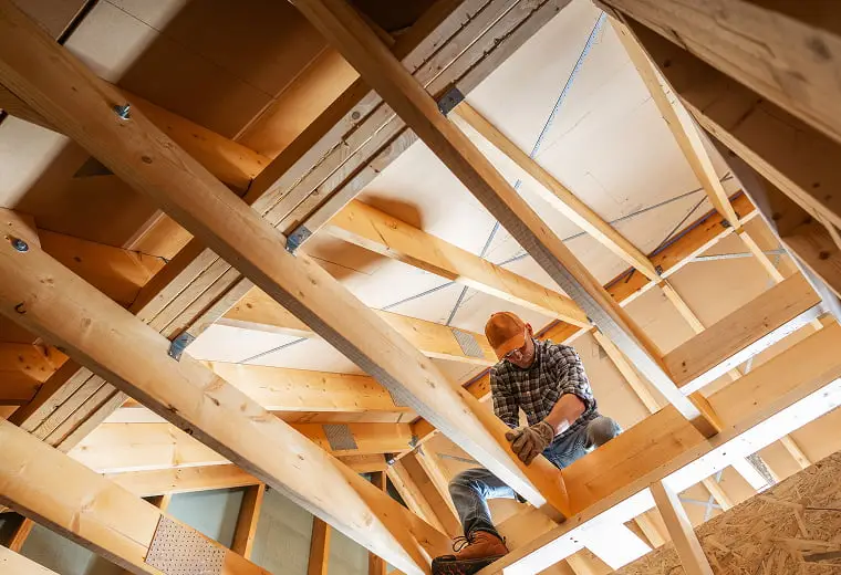 屋根裏で木材の梁を扱う建設作業員。未完成の屋根構造の木製トラスと断熱材に囲まれて作業している様子