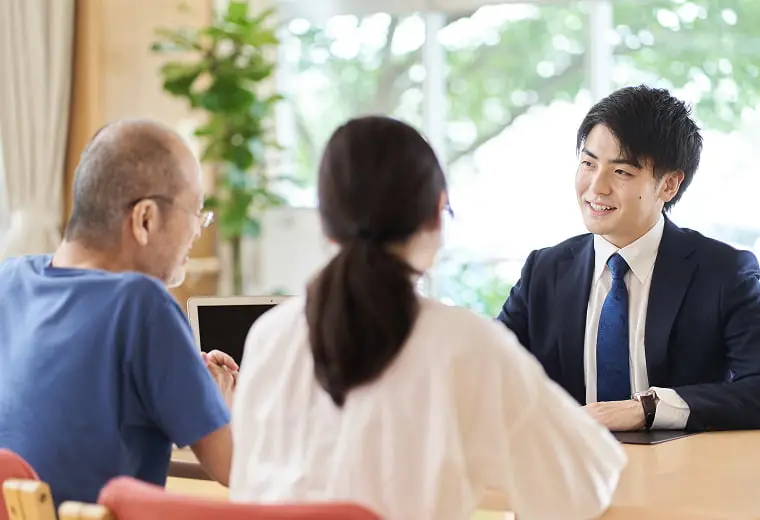 スーツを着た若い男性が、テーブルを挟んで座る年配の男性と女性に笑顔で説明している様子。ビジネスミーティングや顧客との相談風景を示唆する。