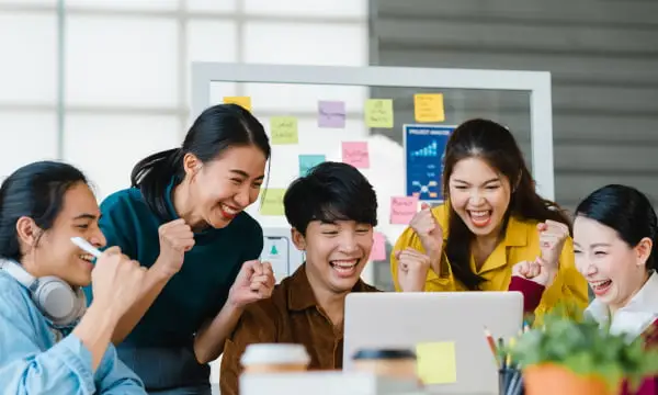 5人のチームメンバーがホワイトボードの前で成功を喜び合っている様子。ホワイトボードには色とりどりの付箋とグラフが貼られ、ラップトップを使って協働作業を行うオフィス環境