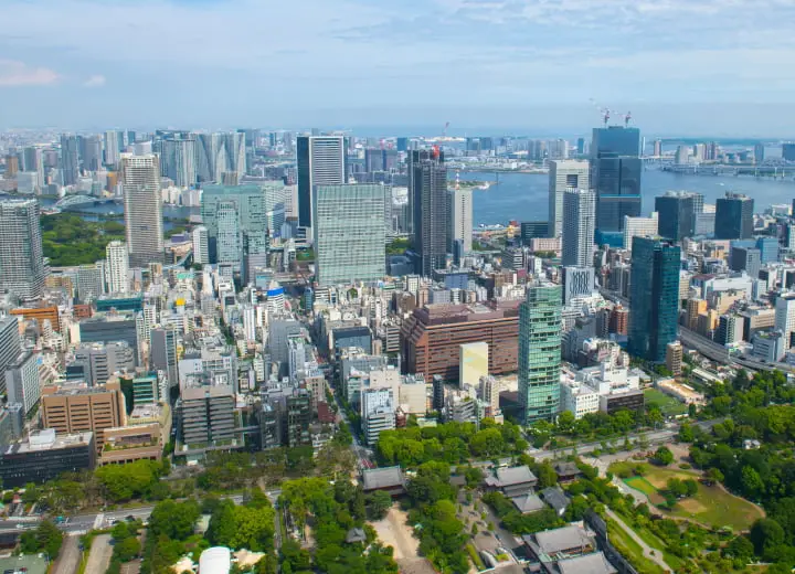 東京の都市景観の空撮。高層ビル群、東京湾、そして緑豊かな公園や伝統的な寺院建築が広がる様子